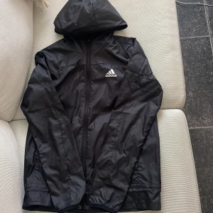 Svart vind/regnjacka från Adidas med huva - Svart  tunn jacka från Adidas med stor vit logga på ryggen och mindre logga på bröstet. Jackan har huva, dragkedja framtill.Perfekt för blåsiga dagar och sportiga outfits. Fungerar till både s/xs storlek 