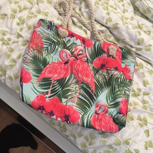 Shoppingväska med flamingomönster - Stor och färgglad shoppingväska med tropiskt motiv av rosa flamingos och gröna blad. Väskan har rejäla rep-handtag i beige och är perfekt för stranden eller city. Materialet känns som bomull eller polyester och mönstret är riktigt somrigt och unikt.