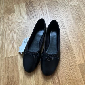 Svarta ballerinaskor med rosett - Klassiska svarta ballerinaskor från H&M i storlek 38. Skorna har rund tå, platt sula och en söt liten rosett framtill. Perfekta att styla med både jeans och klänning för en enkel och clean look.