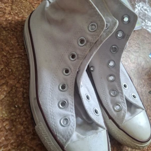 Vita Converse All Star high-top sneakers - Klassiska vita Converse All Star high-top sneakers i canvas med rund tå och platt sula. Skorna har metallfärgade öljetter för snörning och den ikoniska loggan på sidan. Perfekta för en avslappnad och cool streetstil.