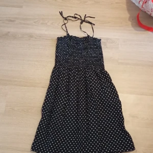 Svart prickig sommarklänning H&M - Supersöt svart kortklänning från H&M med vita prickar och smockad överdel. Klänningen har tunna axelband som knyts upptill och en lätt utställd kjol. Perfekt för varma dagar och ger en lekfull retrovibe.