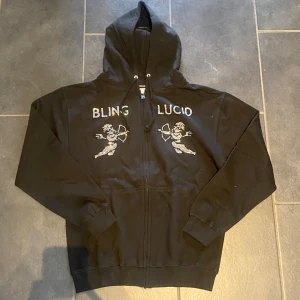Bling Lucid Hoodie Helt Ny!! - Säljer denna svarta Bling Lucid zip-up hoodie med "BLING LUCID" i strass över bröstet, flankerat av två strass-änglar. Det är storlek M. Den är helt ny och aldrig använd. Perfekt för en unik stil med extra bling! Hör mer än gärna av dig vid frågor eller intresse! Skickar gärna fler bilder om det önskas!