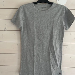 Grå t-shirt från Tommy Hilfiger - En stilren grå t-shirt från Tommy Hilfiger i klassisk modell. T-shirten är kortärmad, har rund halsringning och är tillverkad i mjuk bomull. Liten Hilfiger-logga nertill på framsidan. Perfekt basic-plagg för en clean och enkel look.
