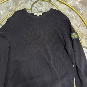 Svart stickad tröja från Stone Island - Svart stickad tröja från Stone Island med klassisk rund hals och ribbad struktur. Tröjan har lång ärm och den ikoniska Stone Island-loggan på vänster ärm. Perfekt för dig som gillar streetwear och vill ha en clean look.