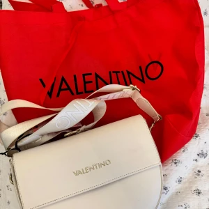 Vit väska från Valentino Bags - Vit crossbody väska från Valentino Bags med guldiga detaljer och bred axelrem med logga. Axelremen har tecken på användning, se bild 🤍