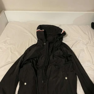 Svart vindjacka från Moncler - Svart vindjacka från Moncler med huva och dragkedja framtill. Jackan har Moncler-logga på ärmen och snygga rödvita detaljer på huvan. Två fickor med tryckknappar och elastiska muddar. Perfekt för dig som vill ha en stilren och sportig. Pris kan diskuteras 