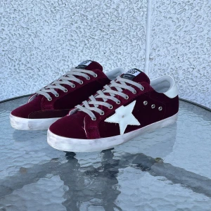 Vinröda sneakers från Golden Goose - Snygga vinröda sneakers från Golden Goose med vit stjärndetalj på sidan, grå snörning och vit hälkappa med logga. Ovandel i sammet och detaljer i skinn. Platt sula och rund tå ger en klassisk men cool vibe.