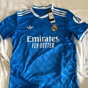 Real Madrid Tredje Ställ 25/26 (#10 Mbappé) - Snygg blå Real Madrid fotbollströja med Mbappé och nummer 10 på ryggen. Adidas logga och klubbmärke på bröstet, vita detaljer och ränder på axlarna. Rund hals och korta ärmar. Materialet är lätt och andas, perfekt för match eller träning.