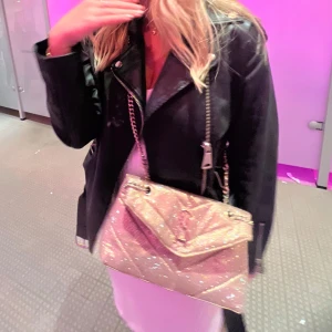 Glittrig axelväska från Steve Madden - Säljer en glittrig axelväska från Steve Madden i guldigt syntetmaterial. Väskan har kedjeremmar med läderdetaljer, quiltad design och magnetlås. Perfekt storlek för det viktigaste och snygg logga framtill. Insidan har fack med dragkedja. Samma modell som den populära YSL väskan! 