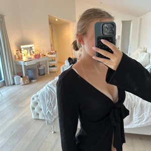 Svart omlottopp med knyt  - Svart långärmad omlottopp från Gina Tricot i storlek M med djup v-ringning och stor knytning framtill. Toppens material är mjukt och stretchigt. Ärmarna är långa och modellen är figurnära men ändå bekväm. Använd kontinuerligt. 