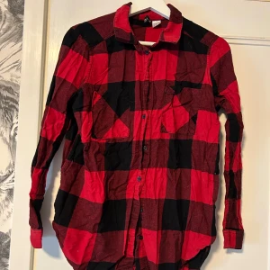 Röd och svart rutig skjorta - Snygg röd och svart rutig längre skjorta med klassisk krage och knäppning framtill. Skjortan har två bröstfickor och långa ärmar. Perfekt för en avslappnad och cool stil.
