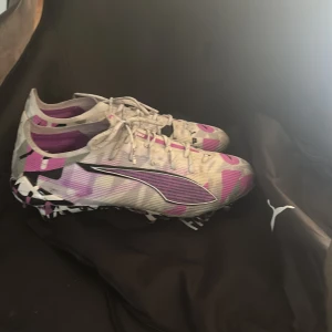 Puma ultra forever ultimate fotbollsskor i vitt och lila - Säljer ett par grymma Puma fotbollsskor med coolt grafiskt mönster i vitt, lila, rosa och svart. Skorna har snörning, låg profil och är tillverkade i syntetmaterial för bästa känsla på planen. Perfekta för dig som vill sticka ut med stil på matchen.Storlek 40,5 men sitter som en 42 då dom är stor i storlek och knappt använda 3 träningar. Ordinarie pris 2600kr