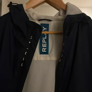 Mörkblå vindjacka från Replay L - Snygg mörkblå vindjacka från Replay i storlek L. Jackan har huva, dragkedja framtill och meshfoder på insidan för extra komfort. Diskret Replay-logga på bröstet och längs dragkedjan. Perfekt för blåsiga dagar och enkel att matcha med allt.