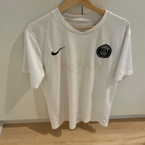 Vit PSG x Balmain fotbollströja - Snygg vit fotbollströja från PSG i samarbete med Balmain och Nike. Tröjan har klassisk rund hals, korta ärmar och svart PSG-logga på bröstet. Nike Swoosh i svart och Balmain Paris tryck i ljus ton på framsidan. Materialet är mjukt och lätt, perfekt för träning eller match.