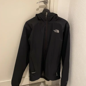 Svart vindjacka från The North Face M - Svart vind/regn jacka från The North Face i storlek M. Jackan har huva, hel dragkedja framtill och meshfoder på insidan. Perfekt för blåsiga eller regniga dagar och enkel att slänga på sig. Snygg och stilren design med logga på bröstet och ryggen.
