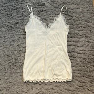 Vit ribbad linnetopp med spets - Supersöt vit linnetopp med tunna axelband och v-ringning dekorerad med spets. Toppen är ribbad och har vågig kant nertill för en extra detalj. Perfekt att styla med jeans eller kjol för en fräsch look.
