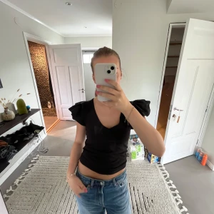 Svart topp med volangärmar - Säljer en svart croppad topp med v-ringning och stora volangärmar. Toppen är figurnära och har en cool, trendig look som passar perfekt till jeans eller kjol. Materialet känns mjukt och stretchigt, troligen bomull eller polyester🩷