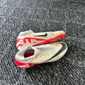 Nike fotbollsskor i vitt och rött - Snygga fotbollsskor från Nike i vitt med röda och svarta detaljer. Skorna har en strumpliknande passform utan klassisk snörning, svart swoosh och röda grafiska mönster på hälen. Yttersulan är röd och vit med fasta dobbar för bra grepp på planen.