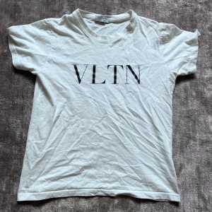 Vit VLTN t-shirt från Valentino - Vit t-shirt från Valentino med svart VLTN-tryck på bröstet. Klassisk rund halsringning och korta ärmar. Tillverkad i mjuk bomull för en skön och avslappnad känsla. Perfekt för dig som gillar stilrena designer och exklusiva märken.
