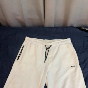 Vita shorts från BOSS - Snygga vita mjukisbyxor från BOSS med svarta dragsnören i midjan och svarta dragkedjor vid fickorna. Byxorna har en relaxed fit och är perfekta för chill dagar. BOSS-loggan är broderad på ena benet för en clean look.