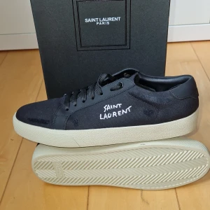 Svarta sneakers från Saint Laurent - Svarta sneakers från Saint Laurent Paris med vit logga broderad på sidan. Skorna har en klassisk låg profil, svarta snören och vit platt sula. Ovandelen är i textil och detaljer i skinn, vilket ger en stilren och lyxig känsla. Allt på bilderna medföljer! Boxen har ett litet jack på ovansidan (bild 4) Storlek 43 passar 42, riktigt feta och extremt trendiga! 🍾