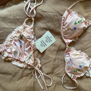 Ljusrosa blommig bikini från H&M - Supersöt ljusrosa bikini från H&M med blommigt mönster och smala knytband. Bikinin har volangdetaljer längs kanterna och är gjord av återvunnen polyester. Perfekt för strandhäng och poolparty!