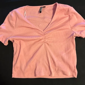 Rosa ribbad croppad topp från Pieces - Superfin rosa croppad topp från Pieces med ribbad struktur och snygg rynkdetalj vid den v-ringade halsen. Kortärmad och i mjukt, stretchigt material som sitter skönt. Perfekt för dig som gillar en enkel men trendig look.