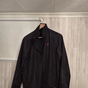 Vindjacka från Ralph Lauren - Svart vindjacka från Ralph Lauren med röd logga på bröstet och röd detalj i nacken. Jackan har dragkedja framtill, hög krage och två fickor med dragkedja. Lätt och smidig modell som passar perfekt till våren, hösten och sommaren. Storleken oklar men ungefär S, passade mig när jag var ca 175. Hör av er vid frågor!