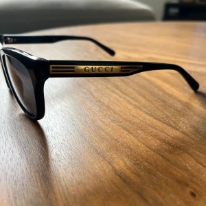 Svarta solglasögon från Gucci - Snygga svarta solglasögon från Gucci med klassisk rektangulär form. Bågarna är i svart med gulddetalj och Gucci-logga på sidorna. Kommer med originalfodral och putsduk. Kanppt använda, som ny!