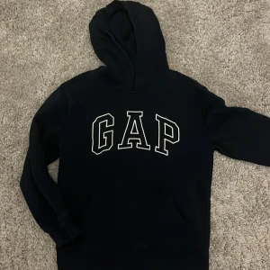 Svart hoodie från GAP Kids - Svart hoodie från GAP Kids med stor vit logga framtill. Klassisk modell med huva och känguruficka. Perfekt för chill dagar eller när du vill ha en enkel och stilren look. Mjuk insida och långärmad design.