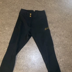 Svarta ridbyxor ZacZess med detaljer - Svarta ridbyxor från ZacZess med snygga guldiga knappar och logga på benet. Byxorna har hög midja, silikonprint för extra grepp och praktiska fickor på sidorna. 