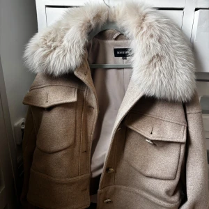 Beige ulljacka med päls från Meotine - Snygg beige jacka från Meotine med stor fluffig fuskpäls-krage. Jackan har stora fickor framtill, knappar och en boxig passform. Perfekt för dig som vill ha en statementjacka med lyxig känsla och coola detaljer.
