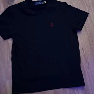 Svart t-shirt från Polo Ralph Lauren - Svart t-shirt från Polo Ralph Lauren med klassisk passform och korta ärmar. Ikonisk röd broderad logga på bröstet. Mjuk och skön bomull som känns riktigt fräsch. Perfekt basic med stilren look och rund hals.