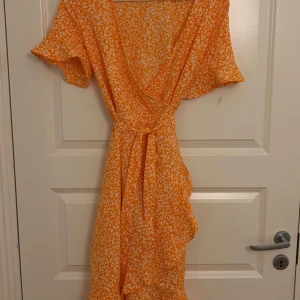 Orange blommig omlottklänning från ONLY - Superfin orange omlottklänning från ONLY med vitt blommigt mönster. Klänningen har korta ärmar, volangdetaljer och knytband i midjan. Perfekt för sommarens alla tillfällen! Luftigt material och snygg v-ringning.Är i storlek M men passar bra på en person med storlek S