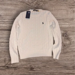 Vit kabelstickad tröja Polo Ralph Lauren - Vit kabelstickad tröja från Polo Ralph Lauren med klassisk rund halsringning och broderad logga på bröstet. Långärmad modell i mjuk bomull, perfekt för lager-på-lager och snygg till jeans eller kjol.