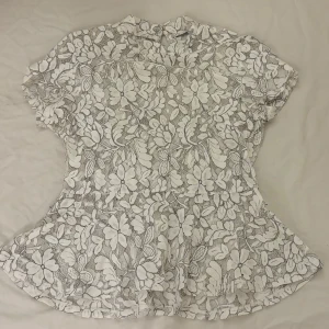 Fin sommartopp i spets - Superfin vit blus i spets med blommor och svarta konturer. Blusen har korta ärmar, liten volang nertill och knäppning i nacken. Perfekt för dig som gillar romantisk och detaljerad stil. Jättefin till sommaren! Går att ha både med ett linne under eller bara en bh. Storleken står inte men skulle säga S.