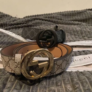 G monogram bälte med spänne - Två snygga G-bälten med klassiskt GG-monogram. Ett har ett matt svart GG-spänne och det andra har ett glänsande guldspänne. Båda bältena är i grått och beige med tydligt GG-mönster och är tillverkade i skinn med canvasdetaljer. Perfekt accessoar för att lyfta din outfit.