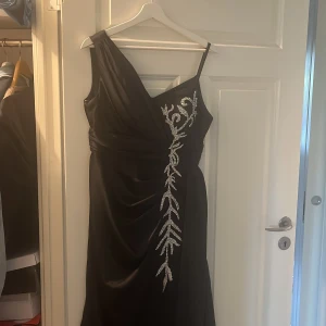 Svart långklänning med silverdetaljer - Snygg svart långklänning med v-ringad omlott upptill och smala axelband. Framsidan har en glittrig silverbroderad detalj som ger extra wow-faktor. Klänningen har en elegant och svepande siluett, perfekt för bal eller fest. Materialet är mjukt och har ett fint fall. Köpt för orginalt för 3000kr.