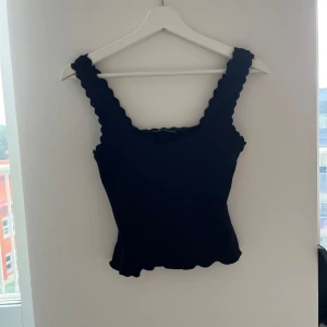 Svart ribbad linnetopp från Zara - Supersnygg svart linnetopp från Zara med breda axelband och vågiga kanter både upptill och nertill. Toppen är ribbad och sitter tight, perfekt att styla med jeans eller kjol. Enkel men ändå unik detalj med de vågiga kanterna.