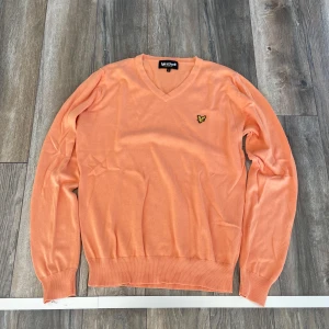 Orange v-ringad tröja från Lyle & Scott - Snygg orange v-ringad tröja från Lyle & Scott med deras klassiska gul fågel-logga på bröstet. Tröjan har långa ärmar och ribbade muddar vid ärmslut och nederkant. Perfekt för dig som gillar stilrena och färgglada plagg.