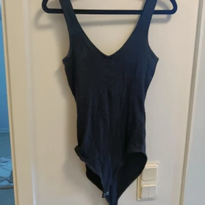 Svart ärmlös bodysuit med djup v-ringning - Svart bodysuit med djup v-ringning och breda axelband. Ärmlös modell som sitter tight mot kroppen och knäpps med tryckknappar i grenen. Perfekt att styla med jeans eller kjol för en clean look.