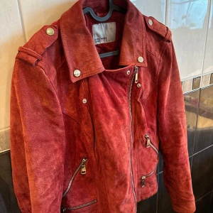 Röd mockajacka från Vero Moda - Snygg röd mockajacka från Vero Moda med cool bikerstil. Jackan har silvriga dragkedjor, axelklaffar och bred krage. Perfekt för dig som vill sticka ut med en statementjacka i höst eller vår. Fodrad och med flera fickor framtill.