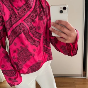 Långärmad rosa bandana mönstrad blus - Säljer en långärmad blus i  skrikrosa med svart paisleymönster och fina detaljer. Blusen har lös passform och är tillverkad i ett mjukt, glansigt material. Perfekt för lite speciellare tillfällen.