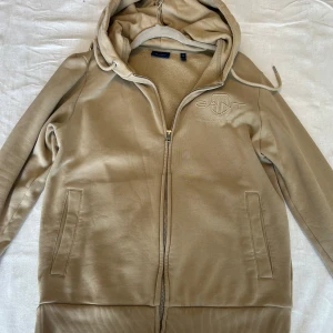 Beige hoodie från GANT med dragkedja - Snygg beige hoodie från GANT med broderad logga på bröstet i storlek M. Nästan helt oanvänd.  Tröjan har huva med snörning, hel dragkedja och två fickor framtill. Insidan är mjukt fodrad för extra komfort perfekt på höst och vinter. Perfekt för chill dagar eller när du vill ha en clean och stilren look. 
