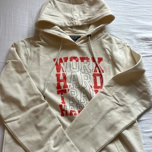 Beige hoodie från OTP med tryck - Snygg beige hoodie från OTP med stor röd och vit text på bröstet: 'WORK HARD TRAIN HARDER'. Tröjan har huva med snörning och en klassisk känguruficka framtill. Perfekt för chill eller gymmet.