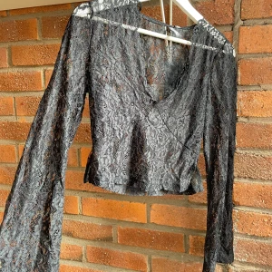 Svart spetsblus från Nelly  - Supersnygg svart blus från Nelly i transparent spets med v-ringning och omlottkänsla. Långa ärmar och croppad passform ger en trendig look. Perfekt för dig som vill sticka ut med en elegant och edgy topp.