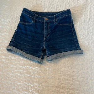 Blå jeansshorts från &Denim - Snygga blå jeansshorts från &Denim med klassisk femficksdesign och uppvikta benslut. Modellen har normal passform och midwaist, med tydliga kontrastsömmar och knapp i metall framtill. Perfekta för sommarens chill dagar.