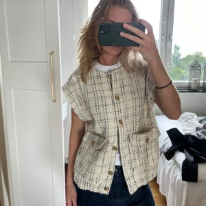 Beige rutig väst med knappar - Säljer en beige väst med svart rutigt mönster och träfärgade knappar framtill. Västen har två fickor med lock och en rak, loose passform. Materialet är grovt vävt och ger en cool, avslappnad vibe. Perfekt att slänga över en t-shirt för en trendig look.
