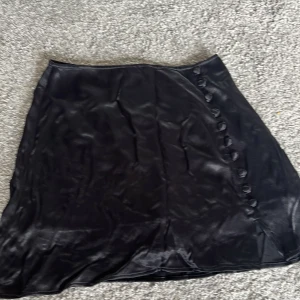 Svart satin kjol från Zara  - svart kortkjol från Zara i glansigt satinmaterial med dekorativa knappar på sidan