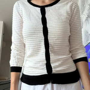 Vit och svart kofta med struktur - Säljer en vit kofta med svart kant och knappar. Koftan har en cool strukturerad yta med små upphöjda ränder och trekvartsärm. Svarta detaljer vid hals, knappslå, ärmslut och nederkant ger en snygg kontrast. Perfekt för dig som gillar stilrena plagg med twist.